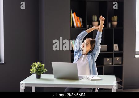 Femme d'affaires qui étire ses bras au-dessus de sa tête et souriant avec plaisir alors qu'elle est assise derrière son bureau de travail Banque D'Images