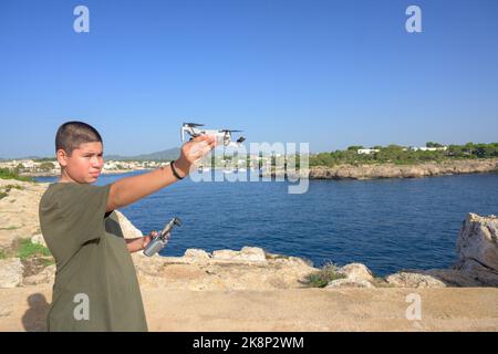 Joyeux adolescent, préparant et finalisant les détails du vol de drone pendant la journée ensoleillée avec la mer méditerranée en arrière-plan Espagne Banque D'Images