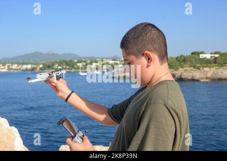 Joyeux adolescent, préparant et finalisant les détails du vol de drone pendant la journée ensoleillée avec la mer méditerranée en arrière-plan Espagne Banque D'Images