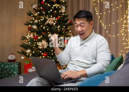 Homme célébrant le nouvel an à la maison et à noël, l'asiatique a obtenu de bonnes nouvelles en ligne à partir d'un ordinateur portable et célébrant la victoire et le succès triomphe assis sur le canapé à la maison près de l'arbre de noël dans le salon. Banque D'Images