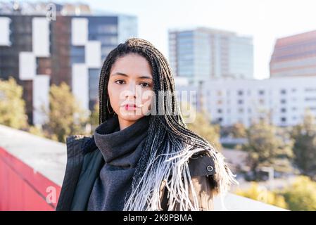 Femme hispanique sérieuse en vêtement d'extérieur chaud avec des dreadlocks regardant la caméra tout en se tenant près de la frontière du bâtiment résidentiel dans la ville Banque D'Images