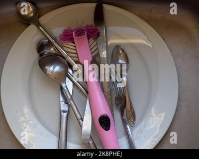 les couverts argentés s'usent dans l'évier avec une plaque blanche et une brosse de nettoyage rose Banque D'Images