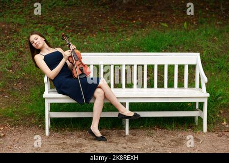 une fille avec un violon est assise sur un banc de parc blanc. Photo de haute qualité Banque D'Images