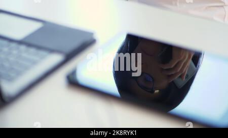 gros plan, sur un bureau blanc, à côté d'un ordinateur portable, le visage d'une femme d'affaires avec des lunettes se reflète sur l'écran de la tablette. elle regarde quelque chose dans un téléphone portable. elle parle sur un téléphone portable. Photo de haute qualité Banque D'Images