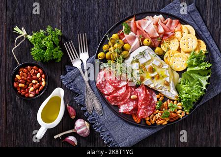 brie salé avec craquelins, salchichon espagnol, saucisses salami, jambon fumé finement tranché, olives vertes, laitue, arachides, pacan sur un plateau noir Banque D'Images