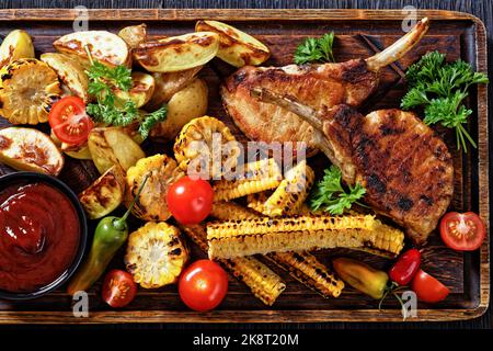 côtelettes de porc tomahawk grillées avec disques de maïs grillés et côtes de maïs, pommes de terre en coin, tomates fraîches et sauce barbecue sur planche à découper rustique, l plat Banque D'Images