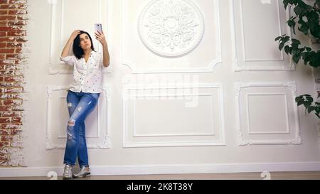 Portrait complet de la jeune femme, fille, brune, en chemise blanche et jeans, debout sur fond de mur blanc avec stuc décoratif, avec smartphone. Regarde l'écran, prend selfie,. Photo de haute qualité Banque D'Images