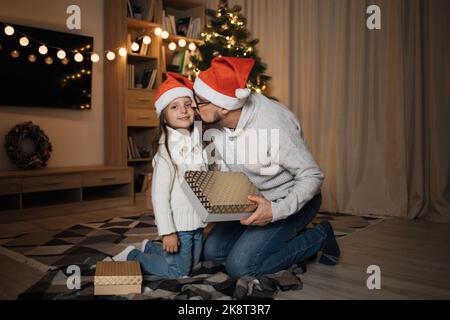 Beau cadeau de partage de mâle caucasien et embrasser sa jolie fille d'âge préscolaire dans le chapeau de santa, assis sur le sol sur le fond de l'arbre décoré, passer des vacances d'hiver heureux à la maison. Banque D'Images