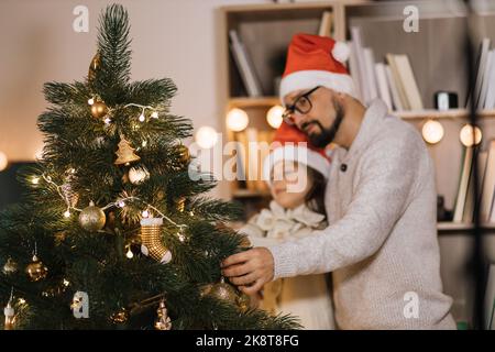 Concentrez-vous sur le jouet de Noël en forme de boule d'or sur l'arbre décoré. Bonne petite fille caucasienne décorant l'arbre avec un jeune père barbu, appréciant se préparer pour la célébration du nouvel an à la maison Banque D'Images