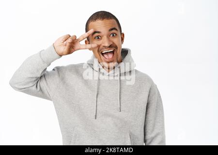 Un gars positif montre le signe de la paix et vous sourit, se tient dans le sweat à capuche gris sur fond blanc Banque D'Images