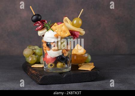 Jarcuterie dans un verre comprend divers types de saucisses, de cornichons, de fromage, de noix, de légumes et de fruits. Version moderne d'en-cas festifs sur fond sombre Banque D'Images