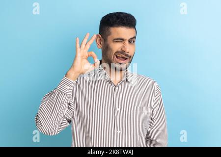 Portrait d'un homme d'affaires barbu ravi debout, regardant l'appareil photo montrant le geste du signe OK, exprimant des émotions positives, portant une chemise rayée. Studio d'intérieur isolé sur fond bleu. Banque D'Images