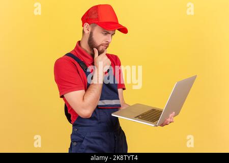 Vue latérale d'un travailleur pensif qui se tient debout et travaille sur un ordinateur portable, qui pense profondément, tient le menton, porte un uniforme bleu et une casquette rouge. Studio d'intérieur isolé sur fond jaune. Banque D'Images