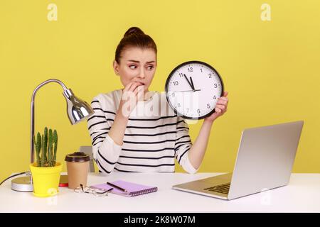 Femme nerveuse malheureuse employée de bureau piquant les ongles, tenant l'horloge murale parlant appel vidéo sur ordinateur portable assis sur le lieu de travail, le temps de se casser. Studio d'intérieur tourné isolé sur fond jaune. Banque D'Images