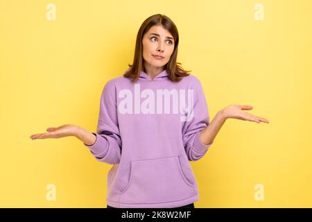 Je ne sais pas. Portrait d'une jeune femme perplexe, debout avec des bras levés et regardant l'appareil photo, portant un pull à capuche violet. Studio d'intérieur isolé sur fond jaune. Banque D'Images