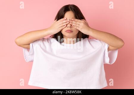 Portrait de triste bouleversé petite fille portant un T-shirt blanc debout couvrant les yeux avec la main, ne veut pas voir à quelque chose de honteux. Studio d'intérieur isolé sur fond rose. Banque D'Images