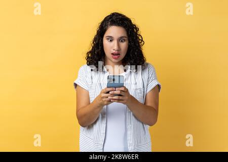 Portrait d'une femme stupéfiante avec des cheveux ondulés sombres utilisant un téléphone mobile et lisant des nouvelles choquantes dans les médias sociaux, regardant l'écran avec de grands yeux. Studio d'intérieur isolé sur fond jaune. Banque D'Images