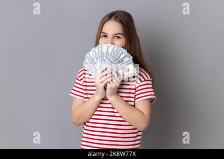 Portrait de charmante petite fille portant un T-shirt rayé cachant la moitié du visage derrière le ventilateur de billets de cent dollars, appréciant l'odeur de l'argent. Prise de vue en studio isolée sur fond gris. Banque D'Images