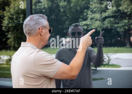 Souriant mûr gris cheveux homme dans des lunettes de soleil stand près du mur de verre du bâtiment, voir la réflexion et le point sur l'espace vide Banque D'Images