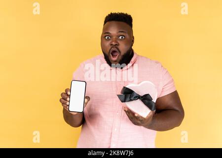Portrait d'un homme barbu surpris portant une chemise rose tenant une boîte en forme de coeur et montrant un smartphone avec écran vide. Studio d'intérieur isolé sur fond jaune. Banque D'Images