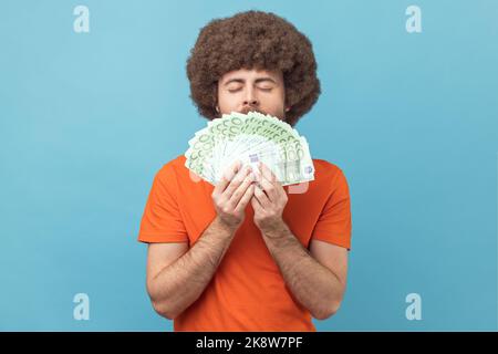 Portrait de l'homme cupide avec une coiffure afro en T-shirt orange qui sent les billets en euros avec plaisir, profitant de gros profits et de la vie riche. Studio d'intérieur isolé sur fond bleu. Banque D'Images