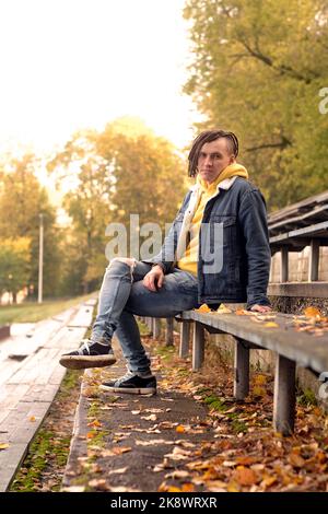 Un jeune homme positif avec des dreadlocks regarde la caméra sur un vieux banc de rue de bleacher en automne doré Banque D'Images