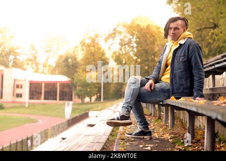 Un jeune homme positif avec des dreadlocks regarde la caméra sur un vieux banc de rue de bleacher en automne doré Banque D'Images