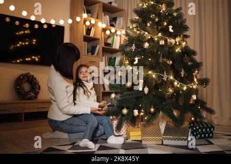 Bonne petite petite école blanche enfant fille décorant arbre de Noël avec la jeune mère heureuse, mettant des jouets sur les branches, appréciant se préparer pour la célébration du nouvel an à la maison, miracle temps concept. Banque D'Images