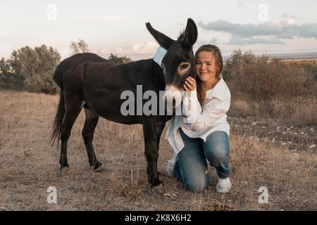 Une fille dans une robe blanche et aérée se dresse avec un âne noir et pose dans la forêt au coucher du soleil, comme dans un conte de fées. Visage bien entretenu avec de beaux cosmétiques. Profitez de la nature et des animaux. Équitation dans la forêt et les montagnes. Photo de haute qualité. Banque D'Images