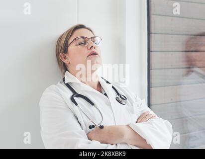 Je ne pense pas que je puisse continuer la journée... une jeune femme médecin attirante qui semble fatiguée tout en se penchant contre un mur dans son bureau. Banque D'Images