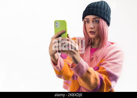 Choqué surpris caucasien adolescent rose-cheveux hipster fille regardant son nouveau smartphone. Gros plan moyen en studio avec arrière-plan blanc. Bonnet noir et gilet rose. Photo de haute qualité Banque D'Images