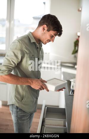 Jeune homme ouvre le réfrigérateur encastré dans sa cuisine. Banque D'Images