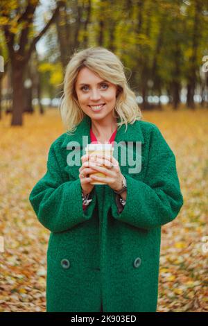 Une femme blonde tient une tasse en papier avec un café à emporter dans ses mains chaudes, debout dans un parc d'automne. Portrait de femme d'automne Banque D'Images
