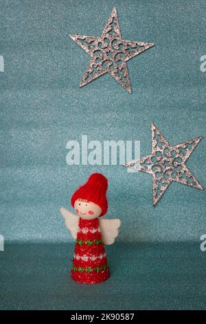 Une jolie figurine d'ange de Noël avec des ailes blanches et des vêtements rouges sur un fond bleu pastel scintillant flou avec deux étoiles argentées brillantes Banque D'Images