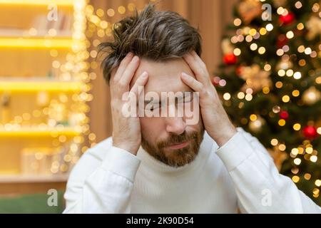 Un homme a un mal de tête pour Noël, une photo en gros plan sur la Saint-Sylvestre près de l'arbre, un homme tenant sa tête avec ses mains, malade, assis sur le canapé seul à la maison. Banque D'Images