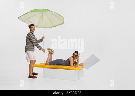 Portrait d'une jeune belle femme dans des vêtements élégants couchés sur un matelas gonflable et homme marin tenant un parapluie isolé sur fond blanc. Banque D'Images