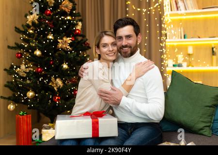 Portrait d'un homme barbu et d'une femme blonde de famille mûre regardant un appareil photo près de l'arbre de Noël, couple célébrant noël et nouvel an avec des cadeaux à la maison assis sur un canapé dans le salon. Banque D'Images