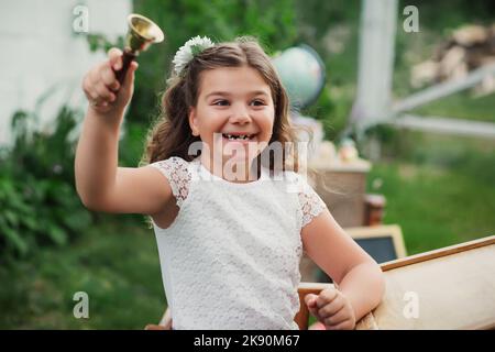 Happy Primary Girl sonne la cloche de l'école et appelle à la première leçon. Une fille intelligente en uniforme de l'école s'assoit au bureau pendant les activités de plein air. Retour à Banque D'Images