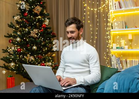 Homme d'affaires prospère travaillant à la maison pour Noël, homme tapant sur ordinateur portable, indépendant travaillant à distance, assis sur un canapé près de l'arbre de Noël célébrant les fêtes du nouvel an. Banque D'Images