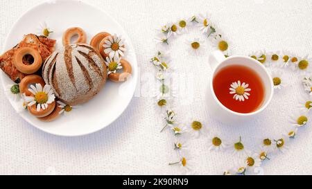 une assiette blanche avec des pâtisseries, des biscuits et des biscuits, décorée de pâquerettes blanches. Sur la soucoupe blanche, il y a une tasse blanche avec du thé de camomille. Autour de lui, il y a un coeur de chamomiles. Photo de haute qualité Banque D'Images