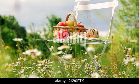Au milieu d'une pelouse de camomille, près d'une forêt, contre un ciel bleu, se dresse une chaise blanche, sur elle une composition d'un panier avec des pommes rouges et du pain, et un bouquet de pâquerettes se dresse. Photo de haute qualité Banque D'Images