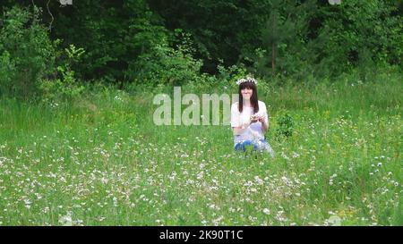 L'été, au milieu d'une pelouse de camomille, dans une forêt, une jeune femme, une brunette tisse une couronne de camomille. Photo de haute qualité Banque D'Images