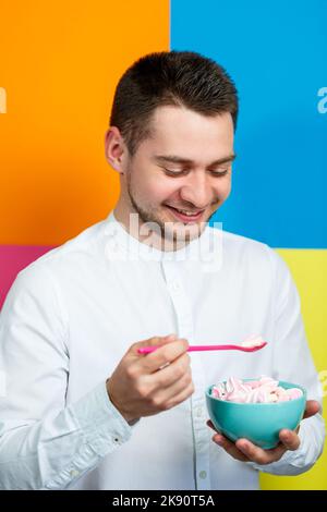 Un homme sur fond jaune, bleu, orange, rose dans une chemise blanche avec un plateau turquoise de guimauves et une cuillère rose. Le gars avec un sourire. Un im Banque D'Images