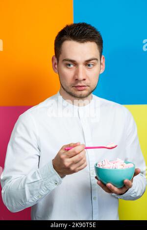 Un homme sur fond jaune, bleu, orange, rose dans une chemise blanche avec un plateau turquoise de guimauves et une cuillère rose. Le gars avec un sourire. Un im Banque D'Images