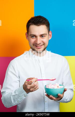 Un homme sur fond jaune, bleu, orange, rose dans une chemise blanche avec un plateau turquoise de guimauves et une cuillère rose. Le gars avec un sourire. Un im Banque D'Images