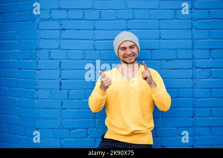 Beau homme avec une barbe et un sourire sur son visage sur le fond d'un mur de briques bleues. Le gars montre des émotions à la caméra, il est dans un jaune Banque D'Images