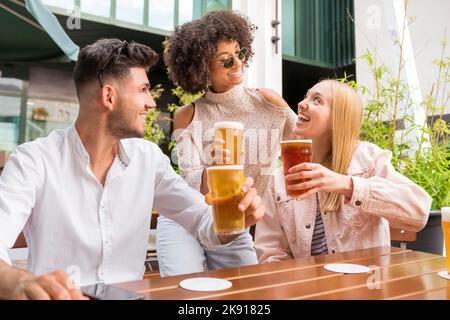 Amis multiraciaux positifs regardant les uns les autres et se clinquant des verres de bière tout en étant assis à table dans le pub extérieur ensemble Banque D'Images