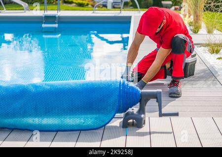 Homme caucasien technicien en travail rouge finition uniforme installation de terrasse composite autour de la piscine extérieure résidentielle. Entretien de l'arrière-cour le Banque D'Images