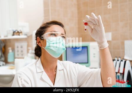 L'assistant de laboratoire tient le tube à essai avec du sang dans ses mains et le regarde. Test sanguin en laboratoire. Scientifique médical en lunettes et masque Banque D'Images