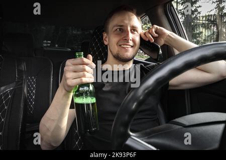 Conducteur souriant ivre avec une bouteille de bière au volant d'une voiture parler sur le smartphone. Concept de conduite avec alcool Banque D'Images